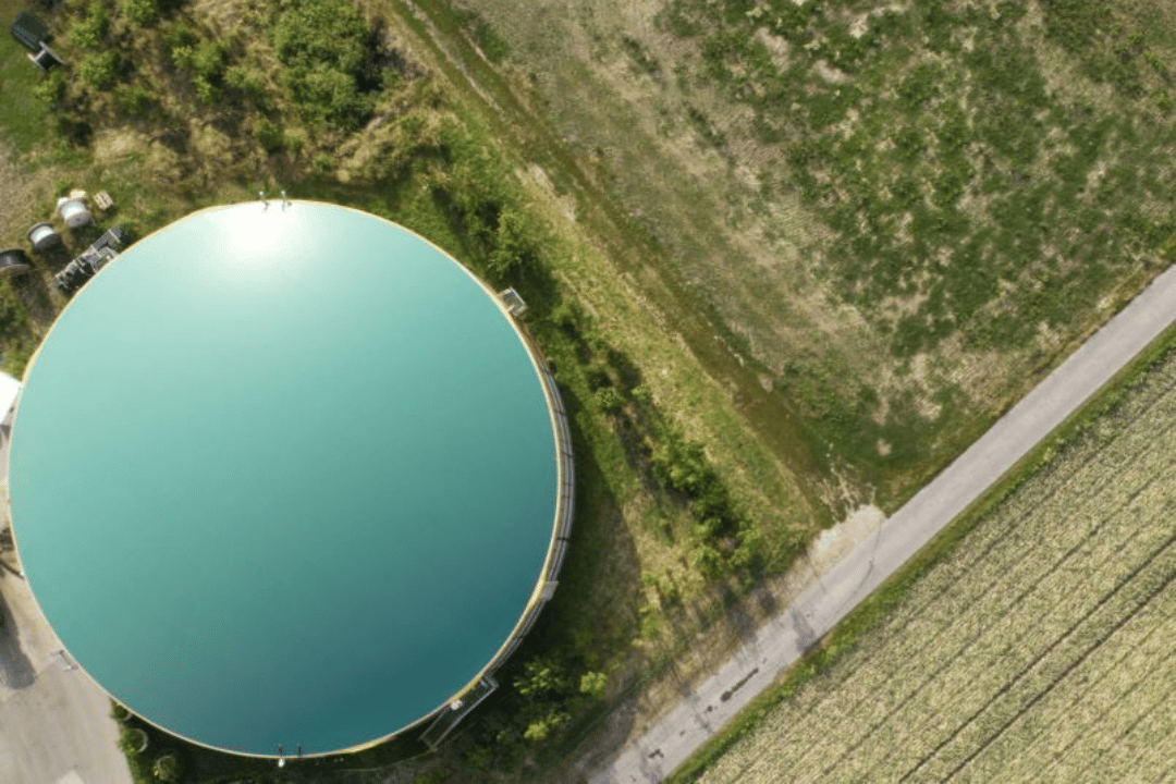 Aerial photograph showing a large circular water tank with a reflective surface surrounded by grassy fields and a paved road. The scene highlights rural infrastructure and agricultural land use with clear contrasts between the water tank, vegetation, and road.