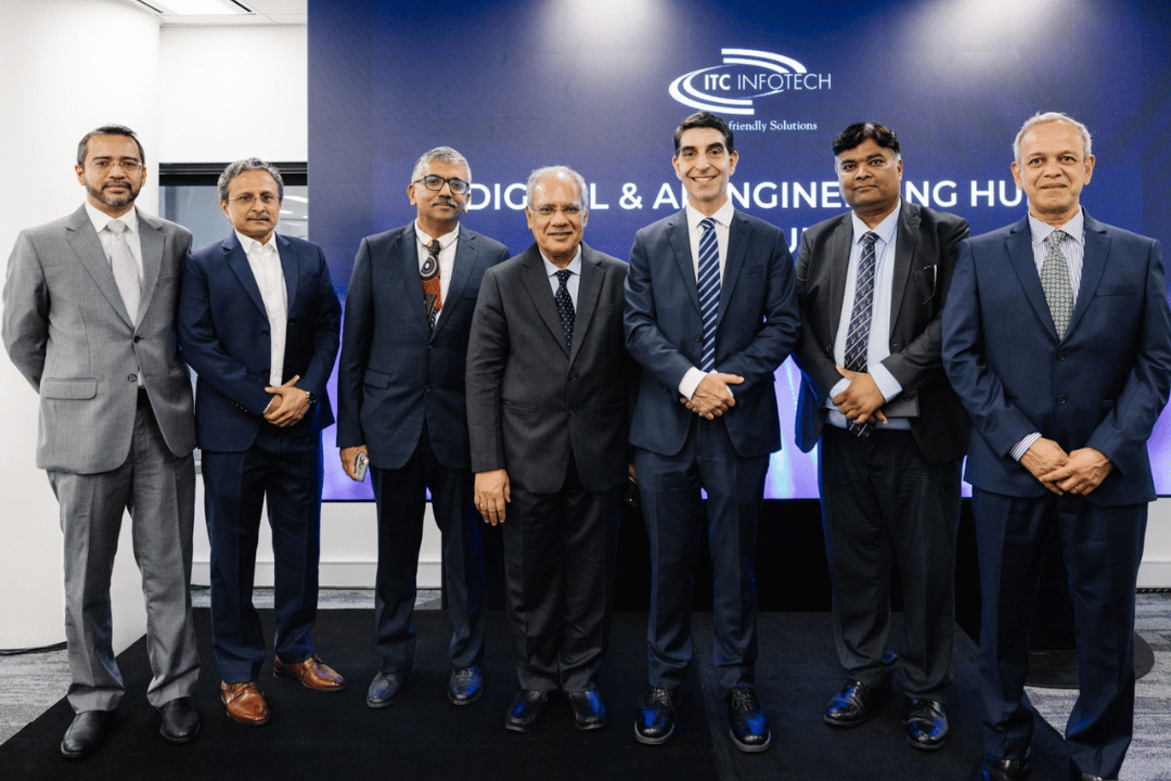 A group of people standing in a row dressed in formal business attire, posing in front of a blue backdrop with "ITC Infotech" and "Digital & AI Engineering Hub" text. The setting appears to be a corporate announcement, highlighting a professional environment.