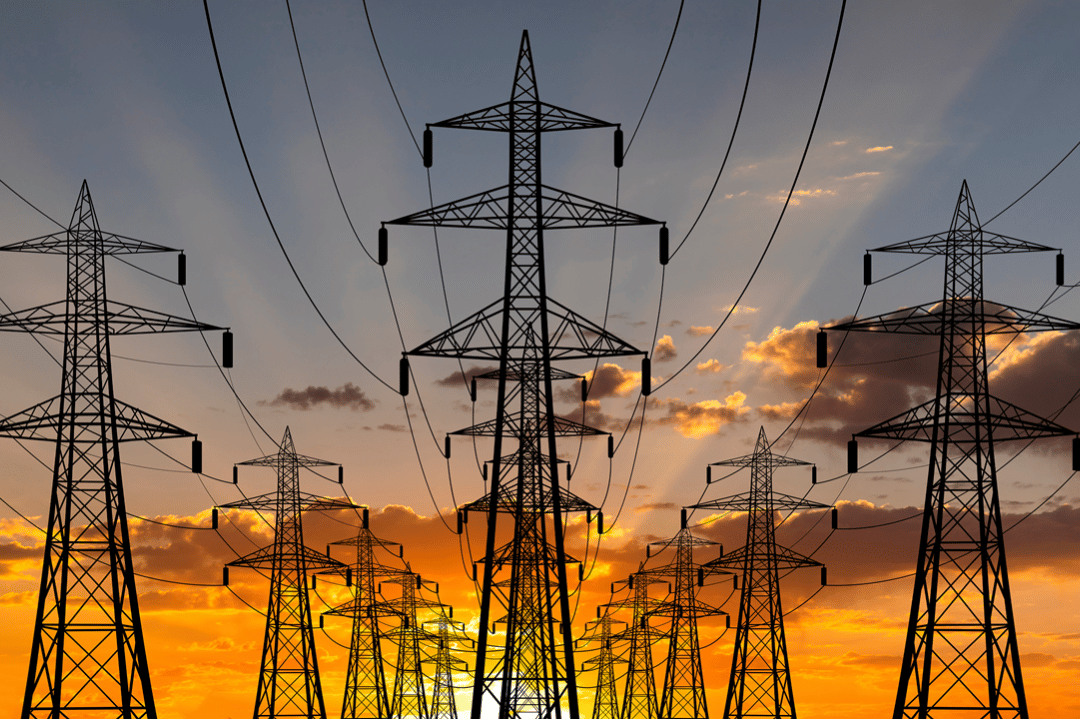 Photograph showing multiple high-voltage power transmission towers silhouetted against a vibrant sunset sky with orange and yellow hues. The towers are connected by numerous power lines, emphasizing infrastructure for electrical energy distribution.