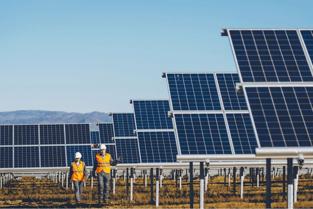 Photograph showing two workers in orange safety vests and white helmets walking through a large solar panel farm under a clear blue sky. Rows of tilted solar panels extend into the distance, highlighting renewable energy infrastructure in a dry, open landscape.
