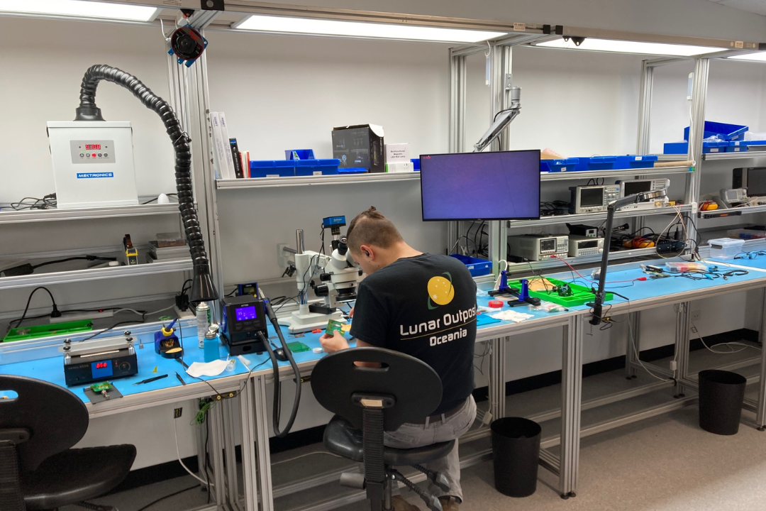 Photograph of a laboratory workspace showing a person seated at a bench working with electronic equipment and tools. The workspace includes multiple monitors, blue work mats, various instruments, and ventilation hoses, indicating a focus on electronics assembly or testing.  