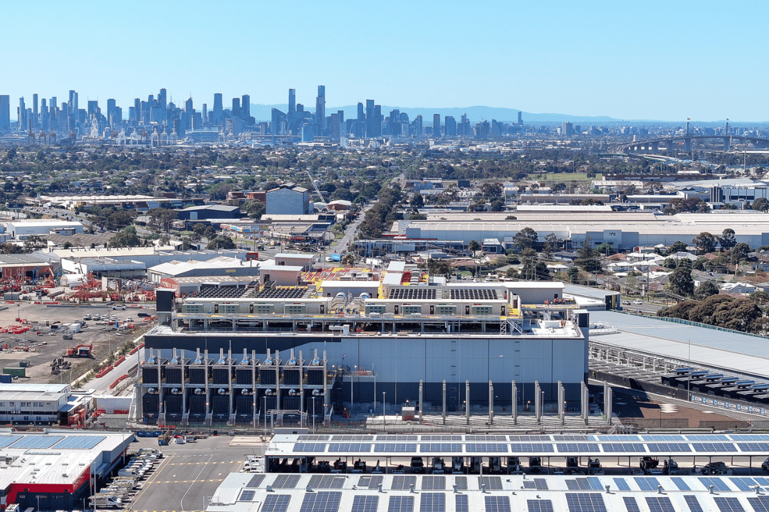 Aerial photograph showing a large industrial warehouse complex in the foreground with a sprawling urban city skyline in the background under a clear blue sky.